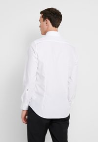 Camicia bianca a maniche lunghe con colletto strutturato, texture liscia, orlo arrotondato e polsini abbottonati, indossata con pantaloni scuri.