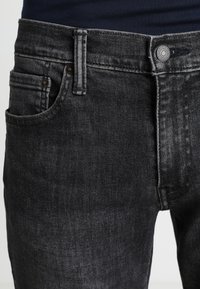 Närbild av midjeområdet på svarta denimjeans, som visar bältesöglor, framficka, knäppning och en del av en marinblå skjorta som är stucken i byxorna.