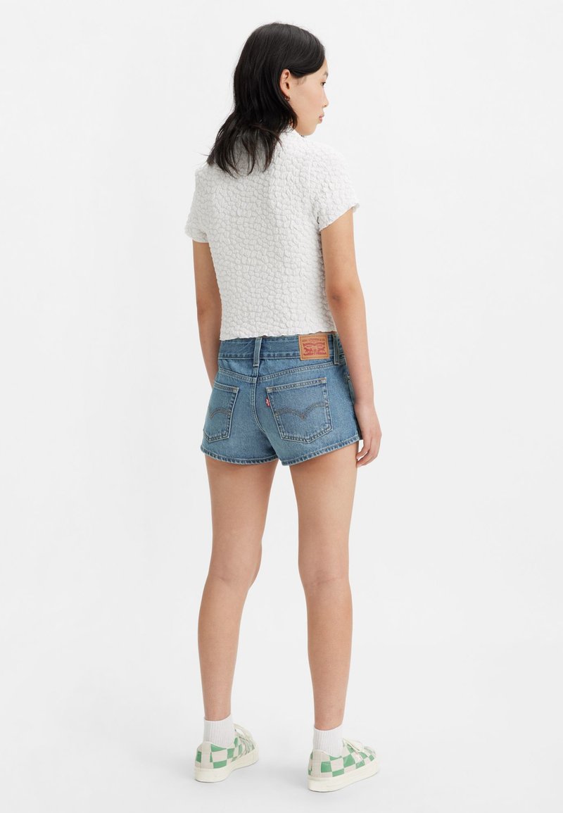 Haut blanc texturé à manches courtes associé à un short en denim bleu taille haute. Baskets blanches avec motif à carreaux verts. Tenue décontractée, vue de dos.
