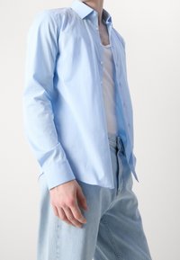 Camisa azul claro abotonada, de tela de algodón, mangas largas, ajuste relajado, con cuello puntiagudo y tapeta de botones visible. Llevada sobre una camiseta sin mangas blanca.