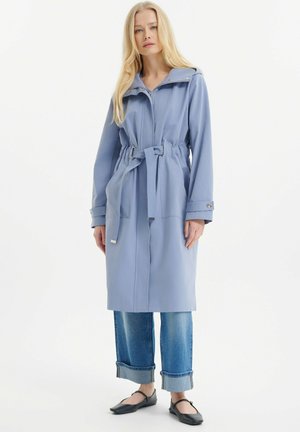 Trench blu chiaro con cintura e cappuccio, con cerniera frontale, bottoni a pressione e maniche con risvolto, abbinato a jeans arrotolati e scarpe basse.