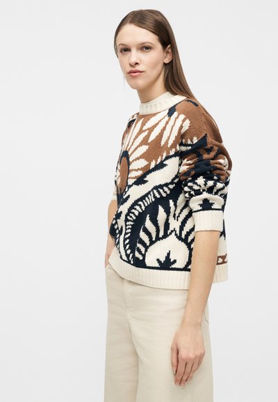 Γυναίκα με ίσια καστανά μαλλιά, που φοράει πουλόβερ με σχέδια σε καφέ, μαύρο και κρεμ χρώμα και παντελόνι κρεμ, στέκεται μπροστά σε απλό φόντο.