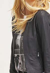 Giacca nera in tessuto strutturato con design aderente, dotata di una cerniera laterale e dettagli in materiali misti, indossata sopra una t-shirt con grafica.