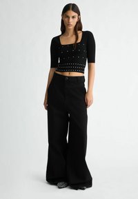 Schwarzes, kurzes Top mit kurzen Ärmeln, verziert mit silbernen Nieten; kombiniert mit weiten schwarzen Hosen. Texturierter Stoff mit einem eleganten Design.