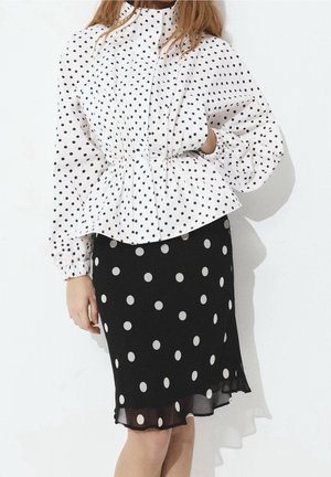 Femme portant un chemisier blanc à manches longues avec des pois noirs et une jupe noire à gros pois blancs.