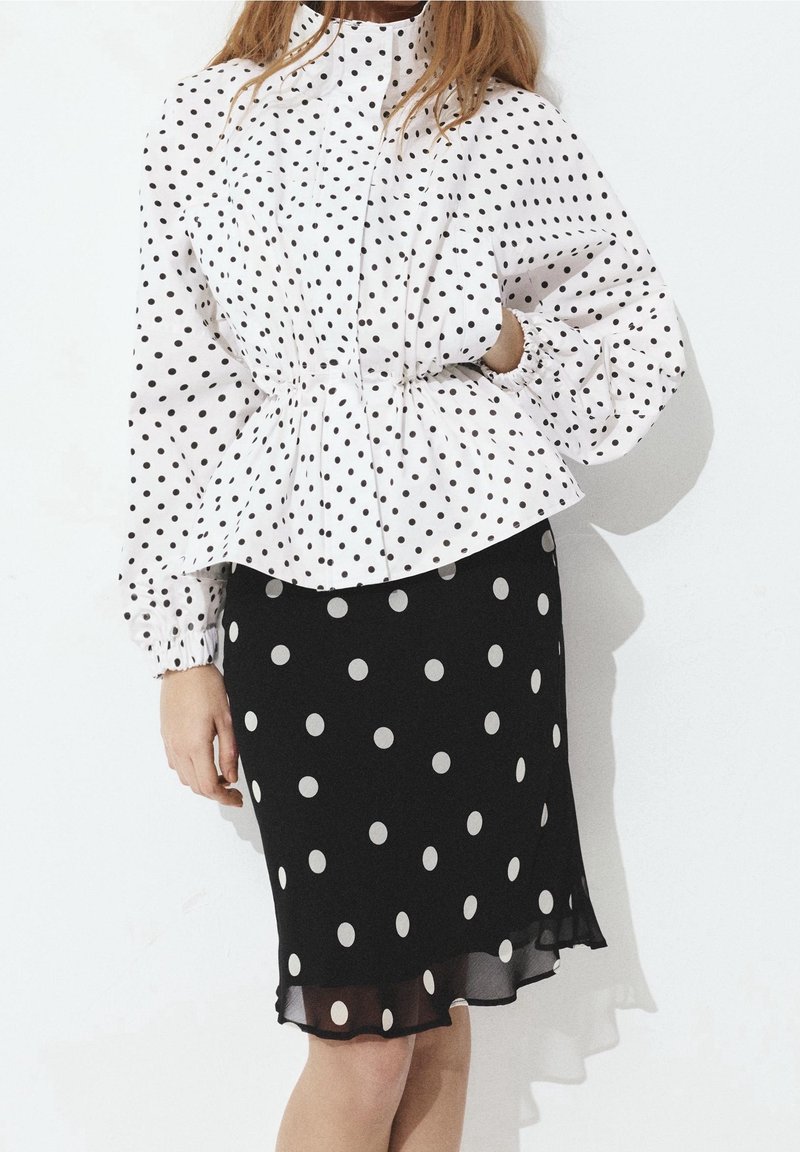 Femme portant un chemisier blanc à manches longues avec des pois noirs et une jupe noire à gros pois blancs.