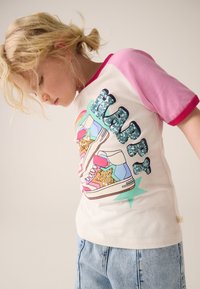 Balta T-krekls ar rozā raglāna piedurknēm, uz kurām redzams dzirkstošs uzraksts "HAPPY" un krāsains sporta apavu attēls ar zvaigznēm, valkāts kopā ar džinsa šortiem.