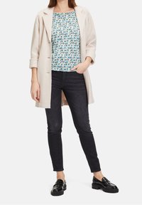 Světlý béžový kabát přes vzorovaný modro-bílý top, kombinovaný s tmavými úzkými džíny a černými mokasíny. Tkanina vypadá hladce a strukturovaně.