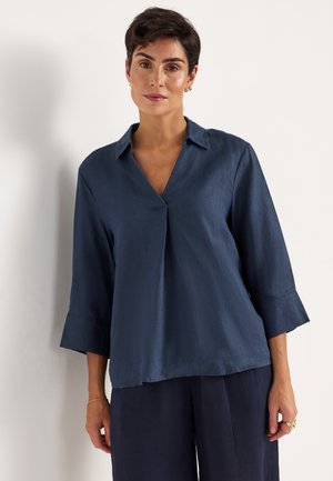 Kvinde med kort mørkt hår iført marineblå skjorte med krave og 3/4 ærmer samt matchende bukser med vide ben foran ensfarvet hvid baggrund.