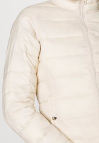 Lys beige pufferjakke med lynlås, ribbet tekstur og vandret syninger. Har sidelommer og høj krave.