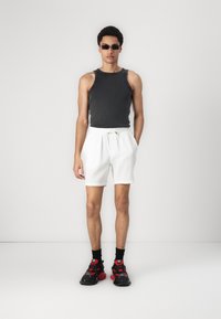 Débardeur sans manches gris, shorts blancs texturés avec cordon de serrage, et baskets noires et rouges. Fond minimaliste.