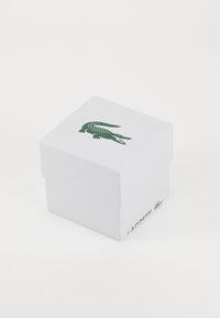 Boîte en carton blanc avec une surface texturée, arborant un logo de crocodile vert sur le couvercle et "LACOSTE" imprimé en vert sur le côté.