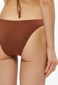 Κάτω μέρος της μέσης και χέρι γυναίκας που φορά καφέ μπικίνι Billabong, φωτογραφημένα από πίσω σε λευκό φόντο.