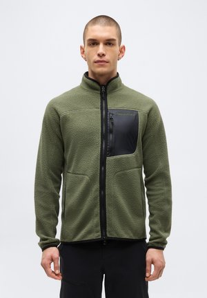 Fiatal férfi olívazöld cipzáras polár pulóvert visel fekete mellzsebbel és fekete nadrággal, egyszínű háttér előtt áll.