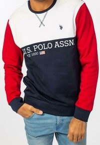 Marinblå, vit och röd långärmad sweatshirt med rundad halsringning. Har texten "U.S. POLO ASSN.", en accent av den amerikanska flaggan och ribbade ärmslut.