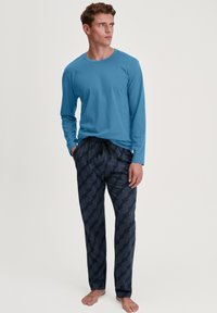 Long-sleeve blue shirt paired with navy patterned pajama pants featuring geometric design and a drawstring waist. Model standing barefoot.
