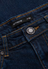 Jean en denim bleu foncé avec couture orange, doté d'un bouton en métal et d'une étiquette noire marquée "ombre M132". Détail usé près du bouton.