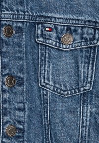 Nära bild av en denimjacka med blått tyg, silverfärgade knappar, en framficka med en triangulär flik och en liten logotyp i röd, vit och blå.