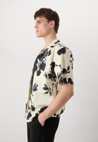 Camicetta a maniche corte di un giallo chiaro con motivo floreale nero, colletto, taschino e texture liscia. Indossata con pantaloni neri, vista di profilo.