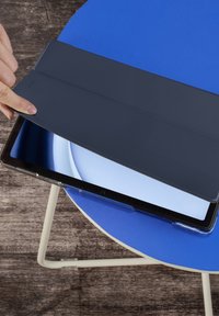 Person hebt den dunkelgrauen Deckel eines Tablets an, das auf einem blauen runden Tisch mit Holzfußboden steht.