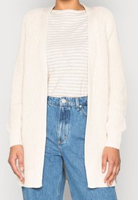 Kreemi neulotusta kardiganista, avoimista etuosista, ribatusta hihasta ja tekstuurista kuviointia, päällä vaalean raidallinen paita ja korkeavyötäröiset farkut.