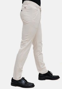 Pantaloni beige in cotone con una vestibilità slim, caratterizzati da una chiusura con bottone, tasche laterali e un'etichetta con logo sulla tasca posteriore. Abbinati a scarpe nere.