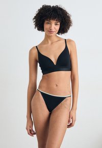 Svart sømløs bralette med dyp V-hals-design og tynne stropper. Matchende svart thong med et kontrasterende elastisk livbånd i hvitt og gult.