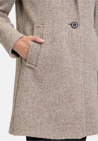 Beige wollenblend jas met een gestructureerde afwerking, voorzien van een enkele zwarte knop en externe zijzakken. Klassieke stijl, middellange design.