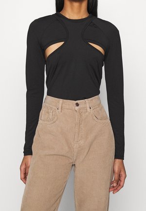 Schwarzes langärmeliges Oberteil mit symmetrischen Ausschnitten unter der Brust, kombiniert mit hoch taillierter beiger Cordhose.