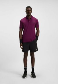 Kurzärmeliges, kastanienrotes Polohemd mit Drei-Knopf-Leiste, kombiniert mit schwarzen Shorts. Schwarze gebrandete Armschützer und Socken, abgerundet mit schwarzen Sneakers.