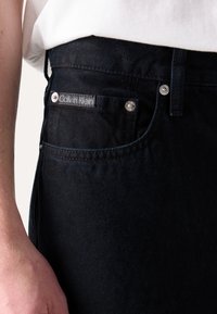 Tamsūs džinsai su Calvin Klein logotipo etikete priekinėje kišenėje, sidabro spalvos detalėmis ir tiesaus kirpimo dizainu.