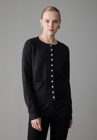 Black cardigan in soft material, featuring a round neckline, long sleeves, front button closure with white buttons, and two front pockets.
