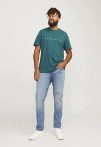 Turkizna majica s belim logotipom, kombinirana s svetlo modrimi kavbojkami in belimi supergami. Model stoji z eno roko v žepu, nevtralno ozadje.