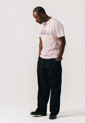 Mand med flettet hår iført en lyserød "Blkvis" t-shirt, sorte vide bukser og sorte sko, står med hænderne i lommerne og kigger ned.