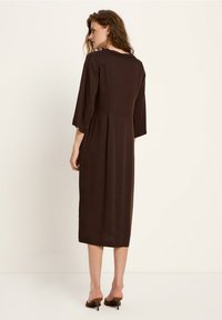 Frau mit welligem Haar, die ein langes braunes Kleid mit Dreiviertelärmeln und braunen High-Heels-Slippern trägt, steht vor einem schlichten hellen Hintergrund.