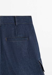 Section de pantalon en denim bleu foncé montrant une poche arrière avec une ouverture horizontale et des passants de ceinture sur fond blanc.