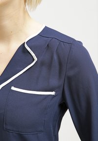 Mörkblå skjorta med vit kant på kragen och en ficka. Lättviktig tyg med en mjuk textur och lätt lyster, med veck på axeln.