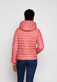 Kobieta w różowej pikowanej kurtce z kapturem i niebieskich dżinsach, pokazana z tyłu na jednolitym jasnym tle.