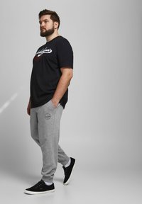 Camiseta de algodón negra con logo blanco, pantalones de chándal de algodón grises con bolsillos laterales y parche de logo, combinados con zapatillas negras y calcetines blancos.