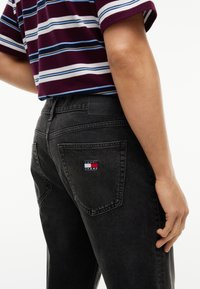 Άτομο που φοράει μαύρα Tommy Jeans με ορατό λογότυπο στην πίσω τσέπη και μπορντό ριγέ κοντομάνικη μπλούζα, στέκεται μπροστά σε λευκό φόντο.