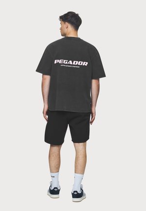 Black oversized T-shirt with "PEGADOR" in pink text on the back, paired with black shorts and navy sneakers, featuring white socks.