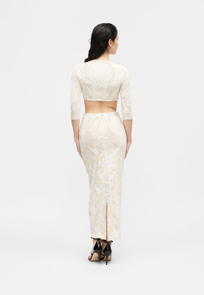 Γυναίκα με μακριά σκούρα μαλλιά που φοράει δύο τεμάχια λευκής και χρυσής με σχέδιο, με κοντό τοπ, φούστα ψηλόμεση και μαύρες ψηλοτάκουνες γόβες.