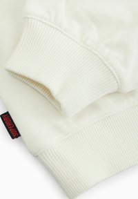 Kremeni dukserica s rebrastim manžetama i rubom. Istaknuta crvena oznaka s brendom "Superdry". Mekana, glatka tekstura kroz cijelu duksericu.