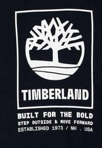 Tessuto nero con una grafica bianca di un albero all'interno di un cerchio. Sotto, il testo recita "TIMBERLAND" e uno slogan in grassetto bianco.