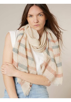 Jonge vrouw met lang bruin haar draagt een zachte beige sjaal met oranje en grijze strepen en een witte mouwloze top.