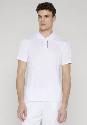 Jonge man die een wit poloshirt met korte mouwen en bijpassende witte broek draagt, staand tegen een effen lichtgrijze achtergrond.