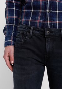 Ciemnoniebieskie dżinsy o dopasowanym kroju, z klasycznym pięciokieszeniowym stylem i subtelną, wyblakłą teksturą, w zestawieniu z niebieską koszulą w kratę.