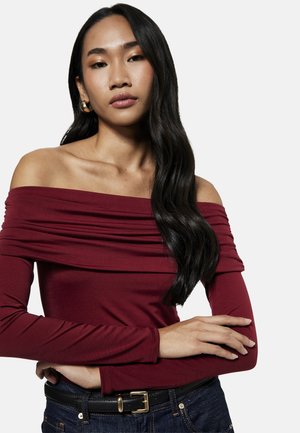 Kobieta z długimi ciemnymi włosami, nosząca złote kolczyki koła, bordową bluzkę z odkrytymi ramionami i długimi rękawami, ciemne jeansy oraz czarny pasek ze złotą klamrą.