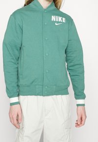 Grön Nike varsityjacka med ståkrage, tryckknappar och vit logotyp. Har sidofickor och ribbade ärmslut med kontrasterande ränder.