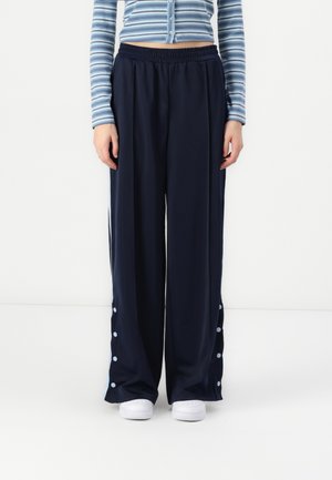 Pantalons larges navy en tissu lisse avec une ceinture élastique et des accents de boutons bleu clair le long des coutures. Portés avec des chaussures blanches.